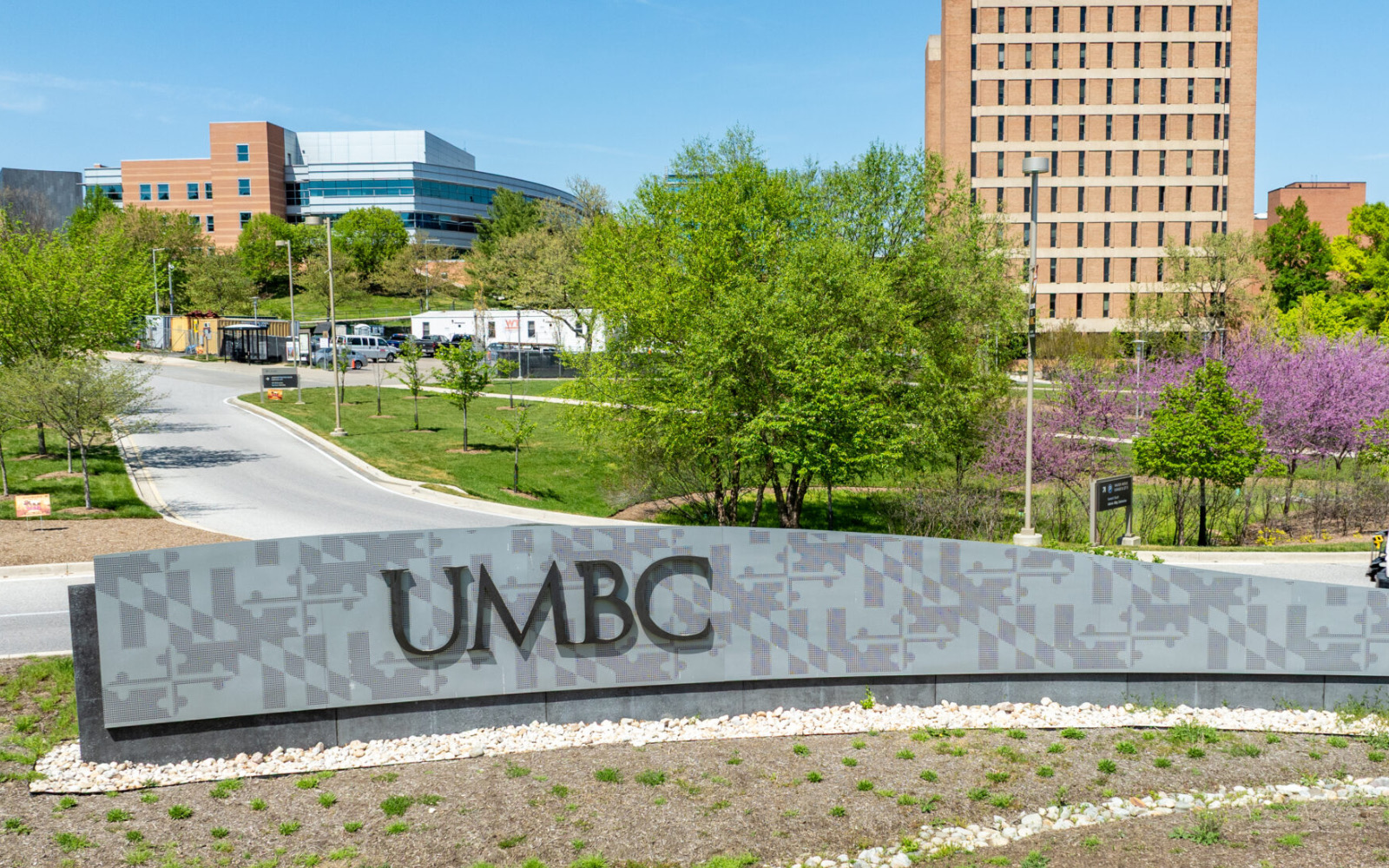 The University of Maryland, Baltimore County entrance sign on a sunny day, with campus buildings and spring trees behind it.